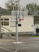 Basketball post and hoop tower, with adjustable mechanism from senior to junior, on an outdoor court with a building and trees in the background